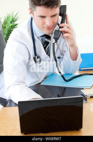 Positive male doctor on phone using his laptop in his office Stock Photo