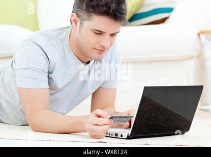 Concentrated man holding a credit card looking at a laptop lying on the floor in the living room Stock Photo