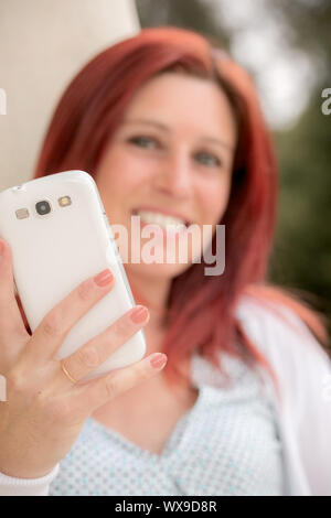 Pretty smiling redheaded woman against an old wall and with her mobile phone on the hand. Focus on her phone Stock Photo