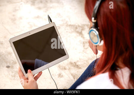 Pretty smiling redhead woman sitting on the floor listening to music or watching a movie on a touch pad. Focus on the tablet Stock Photo