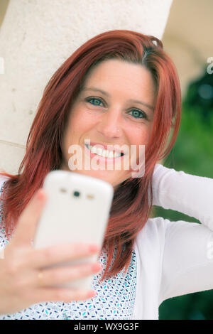 Pretty smiling redheaded woman against an old wall and with her mobile phone on the hand Stock Photo