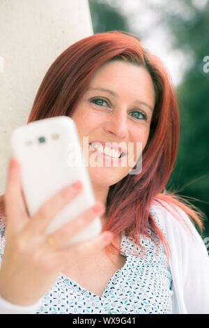 Pretty smiling redheaded woman against an old wall and with her mobile phone on the hand Stock Photo