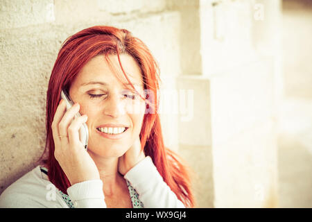 Pretty smiling redheaded woman against an old wall making a call with her cell phone Stock Photo