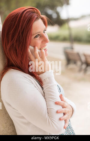 Pretty smiling redheaded woman against an old wall making a call with her cell phone in a park Stock Photo