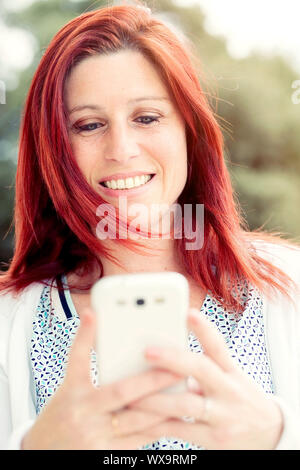 Pretty smiling redhead woman sending messages or make a selfie on her cell phone, against background of summer green park. Stock Photo