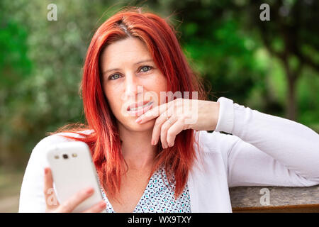 Portrait of smiling beautiful young woman close up with mobile phone, against background of summer green park. Stock Photo