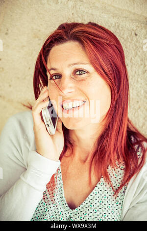Pretty smiling redheaded woman against an old wall making a call with her cell phone Stock Photo