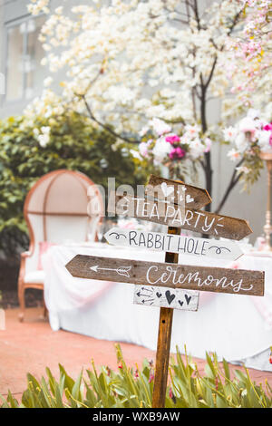 Signpost showing directions for Alice in Wonderland themed wedding during spring with flowering white dogwoods and a banquet table Stock Photo