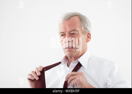 Serious elderly man with a pensive expression putting on his tie holding the two ends in his hands as he stands thinking, head and shoulders studio po Stock Photo