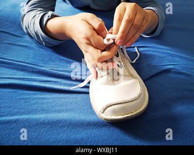 Little boy is learning how to tie shoelaces, close-up on the hands and the shoe on blue textile background Stock Photo