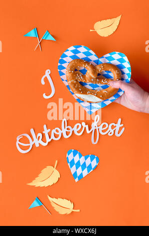 Paper text 'I love Oktoberfest', flat lay on vibrant orange paper background with pretzel in hand, Bavarian blue-white napkins, decorative hearts, fla Stock Photo