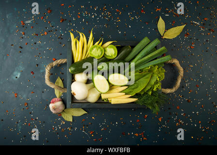 THE VEGETABLES IN THE BOX ON THE STONE A DARK BACKGROUND. YOUNG HERBS ONIONS GARLIC GREEN BEANS YELLOW CORN ZUCCHINI BRIGHT SPIC Stock Photo