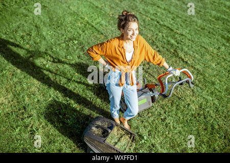 Portrait of a beautiful young woman dressed casually resting while cutting grass with lawn mower on the backyard, view from above Stock Photo