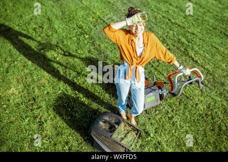 Portrait of a beautiful young woman dressed casually resting while cutting grass with lawn mower on the backyard, view from above Stock Photo