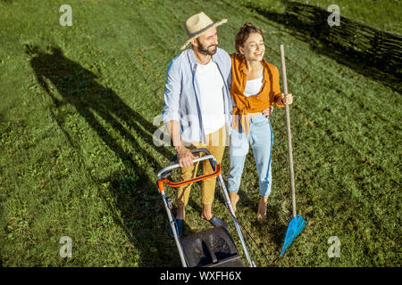 Portrait of a young couple standing together on the green lawn while cleaning backyard with lawn mower and rakes Stock Photo