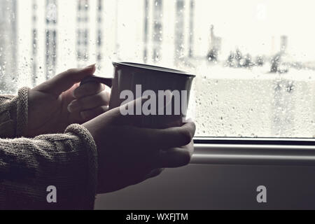Hand holding hot cup of coffee or tea in morning sunlight on a background of wet window with raindrops / comfortable coffee break Stock Photo