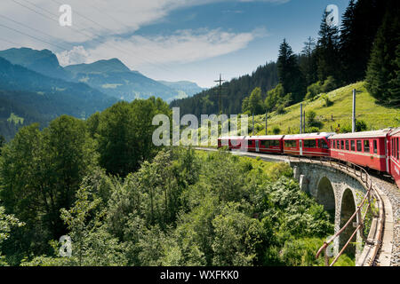 red narrow gauge traincrossing over a stone bridge on a curvy stretch of track in the Swiss Alps Stock Photo