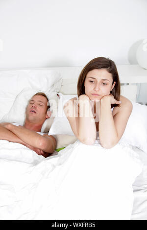 Man lying on his back in bed snoring loudly while his wife, unable to sleep, sits up with a resigned angry expression Stock Photo