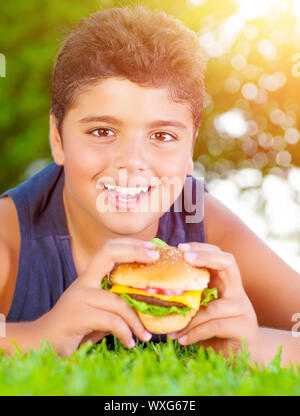 Closeup portrait of cute arabic boy eating burger outdoors, lying down on green grass and enjoying picnic in the park in summer, happy childhood conce Stock Photo