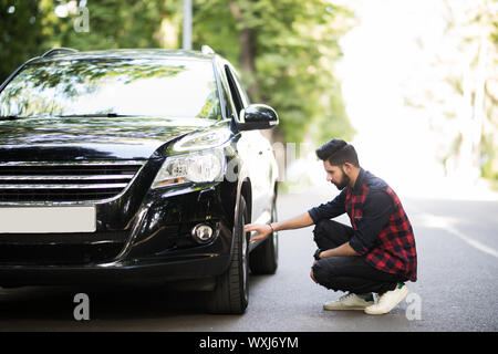 Cheerful young businessman has problems with the wheel of his car. He is kneeing and looking at it with seriousness Stock Photo