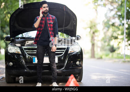 A young man with a silver car that broke down on the road.He has set up a warning triangle.He is waiting for the technician to arrive. Stock Photo