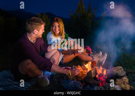 Couple cheering, rosting sausages on the campfire, drinking beer and relaxing by the campfire in the mountain top in the night. Romantic campfire conc Stock Photo