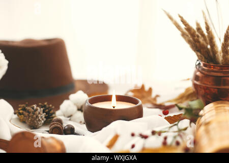 Hygge lifestyle. Candle, berries, fall leaves, herbs, acorns, nuts and brown hat on white fabric. Autumn mood. Hello autumn, cozy inspirational image. Stock Photo