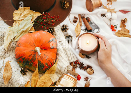 Pumpkin and candle in hand with berries, fall leaves, anise,herbs, acorns, nuts, cinnamon, cotton on white knitted sweater. Hygge lifestyle, cozy autu Stock Photo