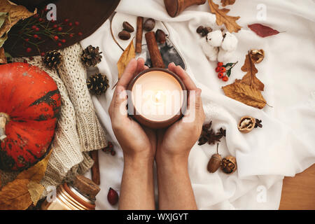 Hygge lifestyle, cozy autumn mood. Pumpkin and candle in hand with berries, fall leaves, anise,herbs, acorns, nuts, cinnamon, cotton on white knitted Stock Photo
