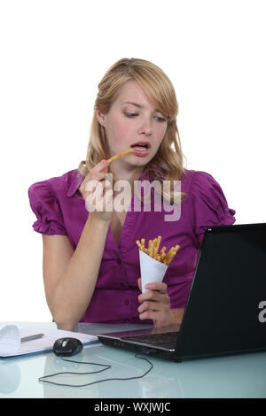 Woman eating French fries at desk Stock Photo