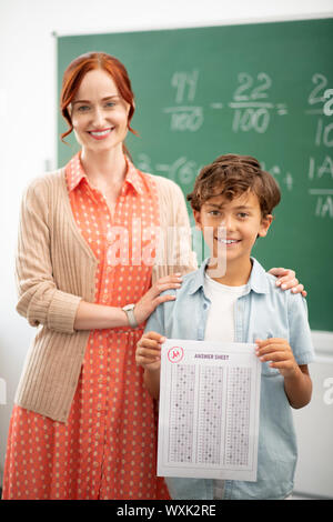 Dark-eyed pupil smiling while standing near teacher Stock Photo