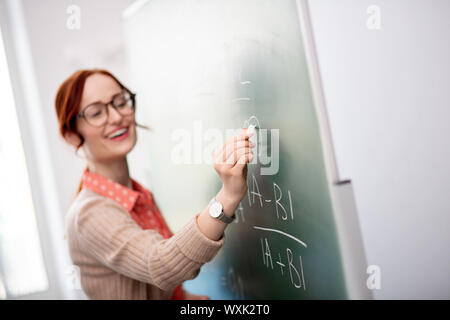 Teacher wearing glasses holding chalk while writing on blackboard Stock Photo