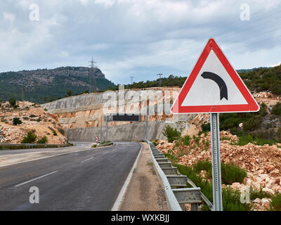 Asphalt road with road sign dangerous curve and metal arch with blank banner. Stock Photo