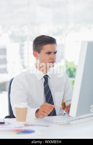 Young businessman frowning while looking at computer and eating Stock Photo