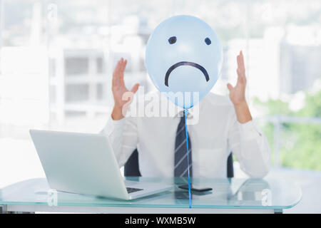 Blue balloon with sad face hiding angry businessmans face in bright office Stock Photo