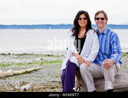 Interracial couple in forties together on beach, sitting on log Stock Photo