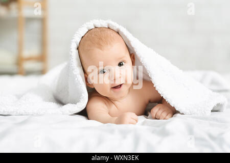 Adorable smiling baby boy lying on bed under white blanket Stock Photo