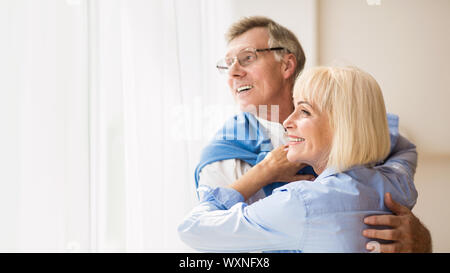 Happy senior couple looking through window and hugging Stock Photo