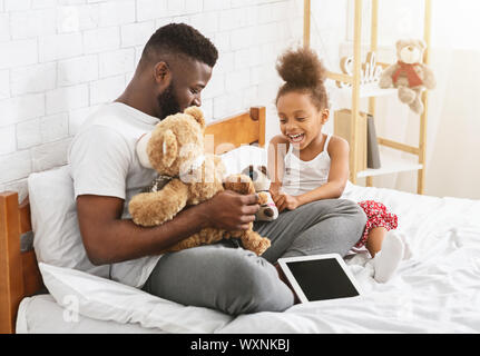 Cheerful afro girl playing toys with her daddy, sitting on bed