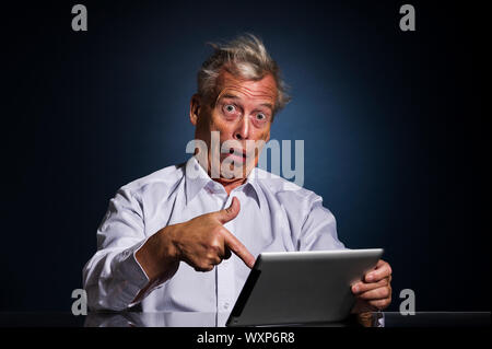 Shocked senior man pointing to his tablet computer with his finger with a look of appalled disbelief and confusion, humorous upper body studio portrai Stock Photo