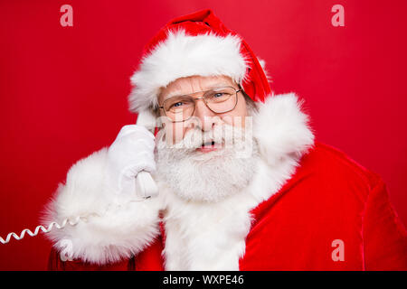 Stylish Saint Nicholas with white beard in eyeglasses costume headwear answer talk on the phone isolated on red background look at camera Stock Photo