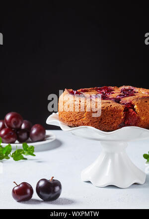 famous plum torte on a white cake stand with fresh plums on a table with black background, copy space Stock Photo