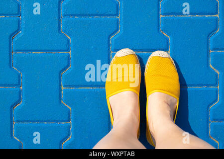 Female legs in yellow shoes or moccasins on a blue tile on a sunny day.