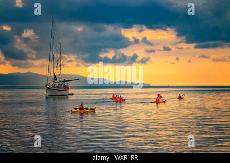 Yacht in the mediterranean sea at sunrise, Cagliari, Italy. People sail on kayaks on the open sea under the sun. The sun illuminates the island with Stock Photo