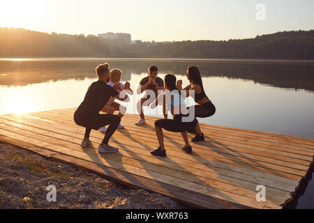 A group of sports people do squat exercises in a park by the lake Stock Photo