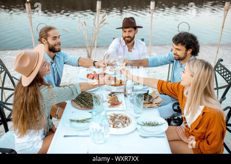Group of young friends having fun, clinking wine glasses during a festive dinner at the beautifully decorated table near the lake outdoors Stock Photo
