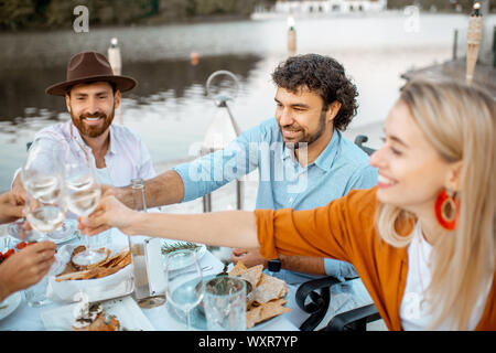 Group of young friends having fun, clinking wine glasses during a festive dinner at the beautifully decorated table near the lake outdoors Stock Photo