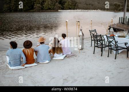 Group of young friends sitting on the beach, enjoying evening time together during a festive meeting and dinner outdoors Stock Photo