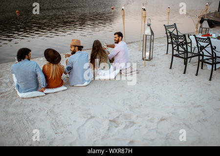 Group of young friends sitting on the beach, enjoying evening time together during a festive meeting and dinner outdoors Stock Photo