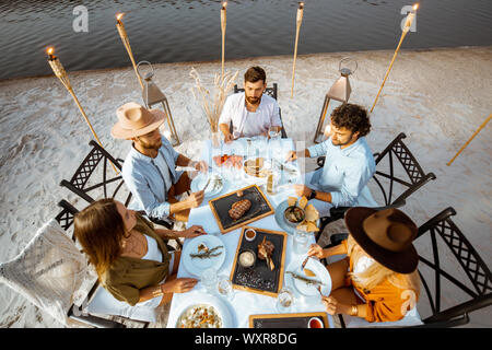 Group of friends having a festive dinner at the beautifully decorated table with delicious meals on the beach at dusk Stock Photo